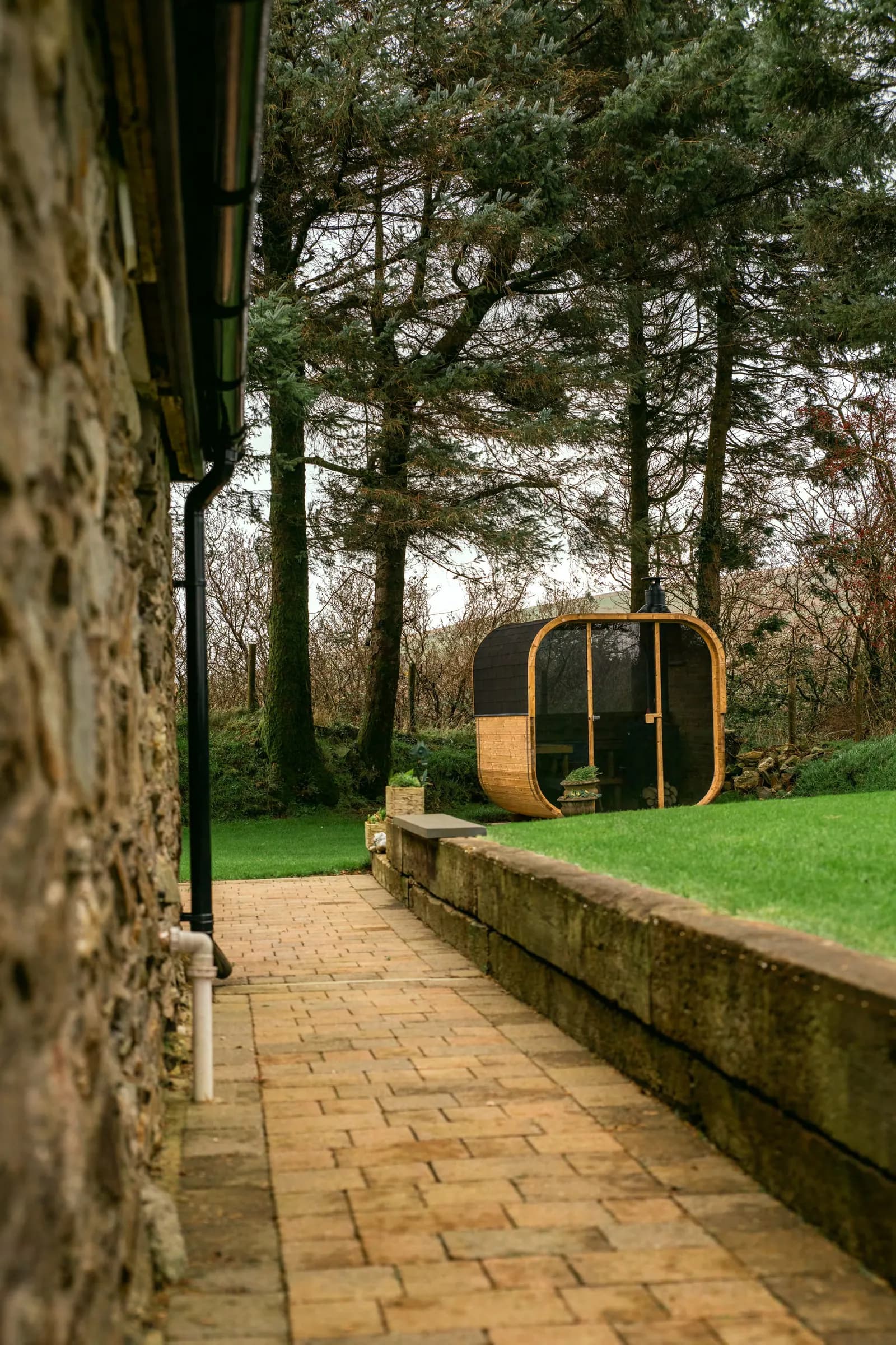 A Hekla cube sauna standing on a stone patio beside a lawn and trees